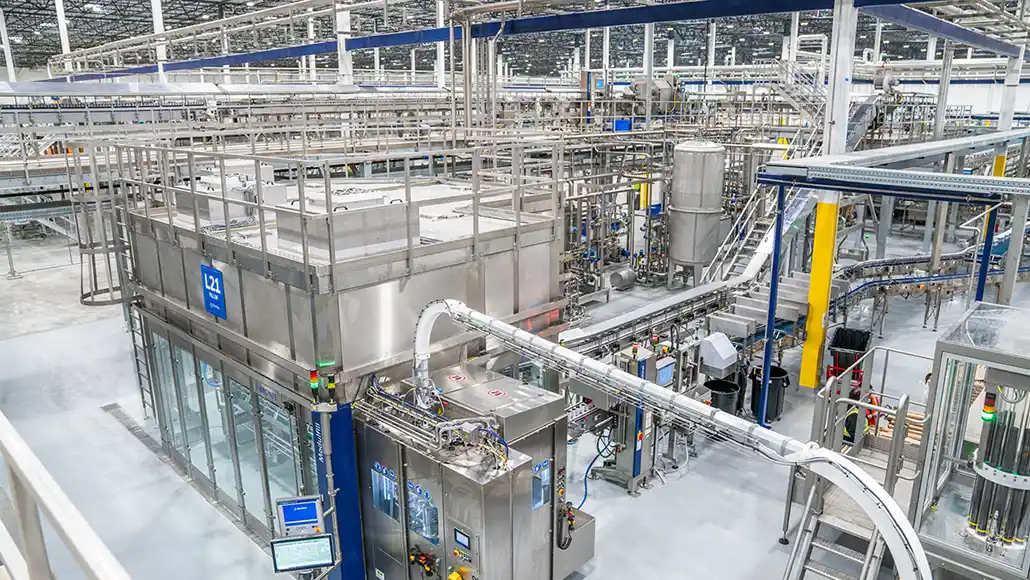 Industrial bottling/processing plant with stainless steel equipment, tanks, and overhead conveyors in a clean factory setting.